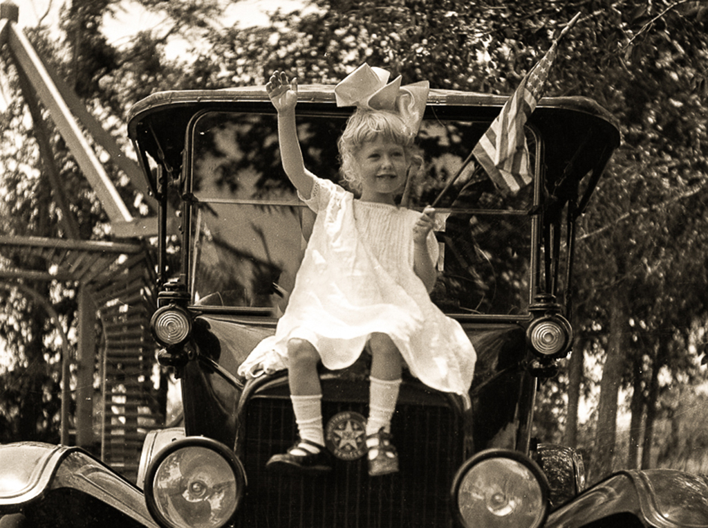 Little girl sitting on a car waving a tiny american flag. circa 1910-1925.